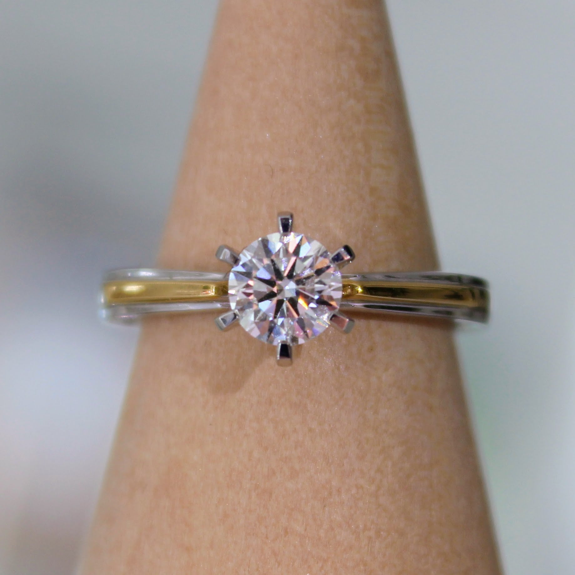 A close-up of a two-tone diamond engagement ring displayed on a wooden ring holder, featuring a brilliant solitaire diamond set in 18K solid gold. The perfect blend of luxury and modern design.