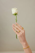 Hand mit Drei-Stein-Verlobungsring aus 18K Gold und 3,50 ct Labordiamanten hält eine weiße Rose; dazu passender Ring und Armband