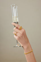 Hand mit Fancy Yellow Statement-Ring aus 18K Gold und 4,00 ct Labordiamanten hält ein Champagnerglas; dazu passendes Diamantarmband